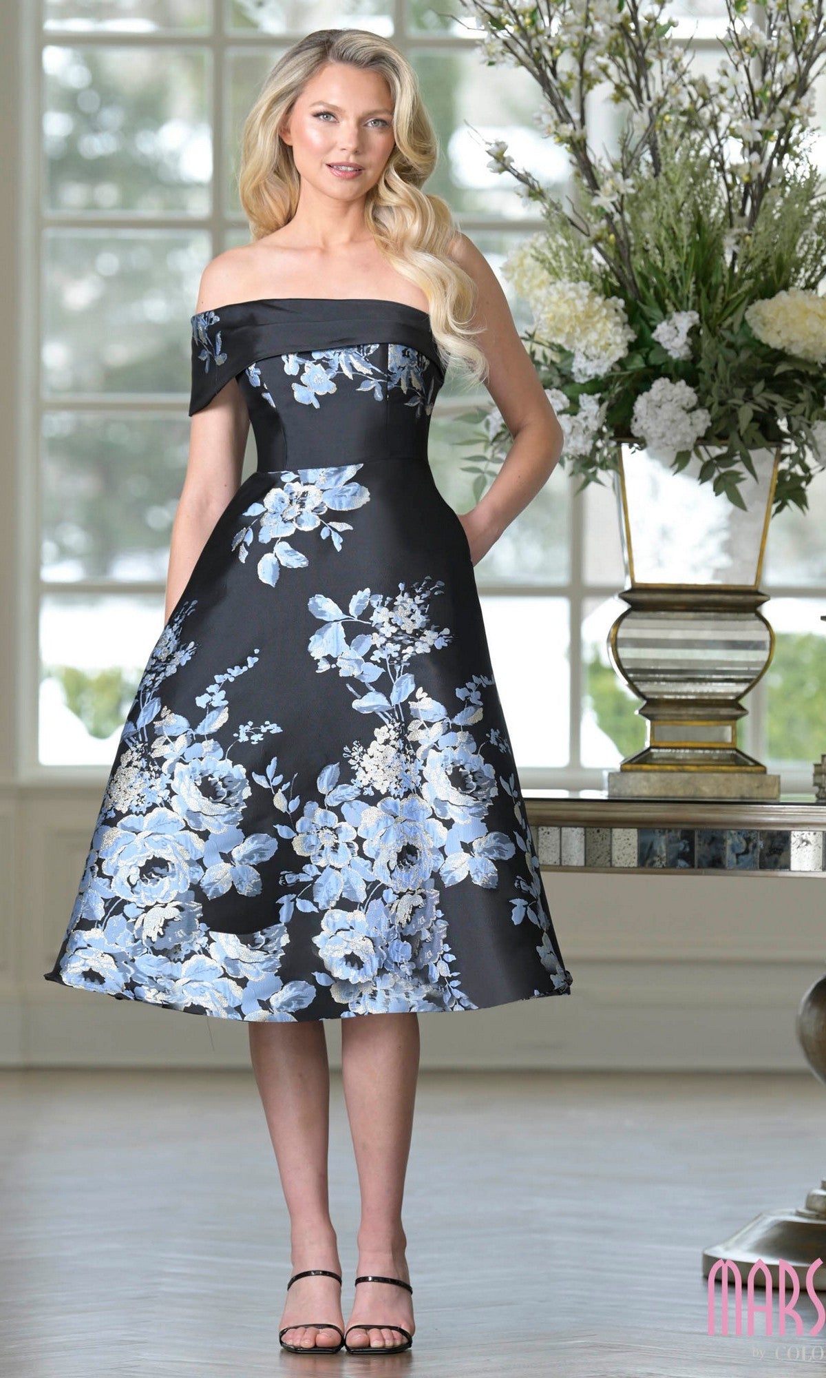 A woman in the Marsoni MV1415 knee-length semi-formal dress in Black Multi stands indoors beside a large flower vase.